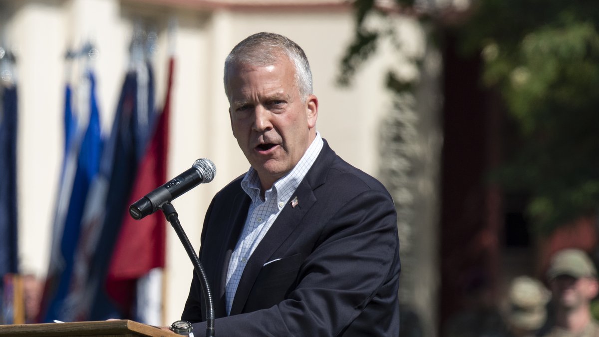 Alaska Sen. Dan Sullivan in a navy blazer speaking into a microphone at a wooden podium outdoors with colorful flags and soldiers behind him