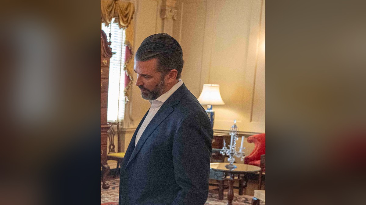 Donald Trump Jr. in a navy blazer and open-collar white shirt looking down inside a formal State Department room