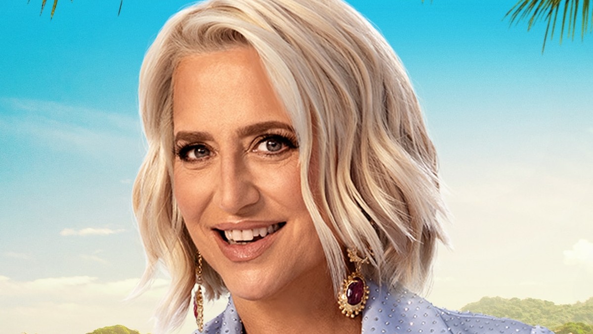 Dorinda Medley smiling with wavy platinum hair and jeweled earrings in front of a tropical beach