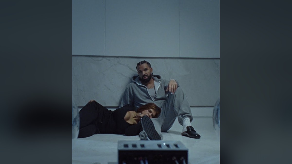 Drake in a gray hoodie sitting on a studio floor beside a woman in a black top during the Janice STFU video