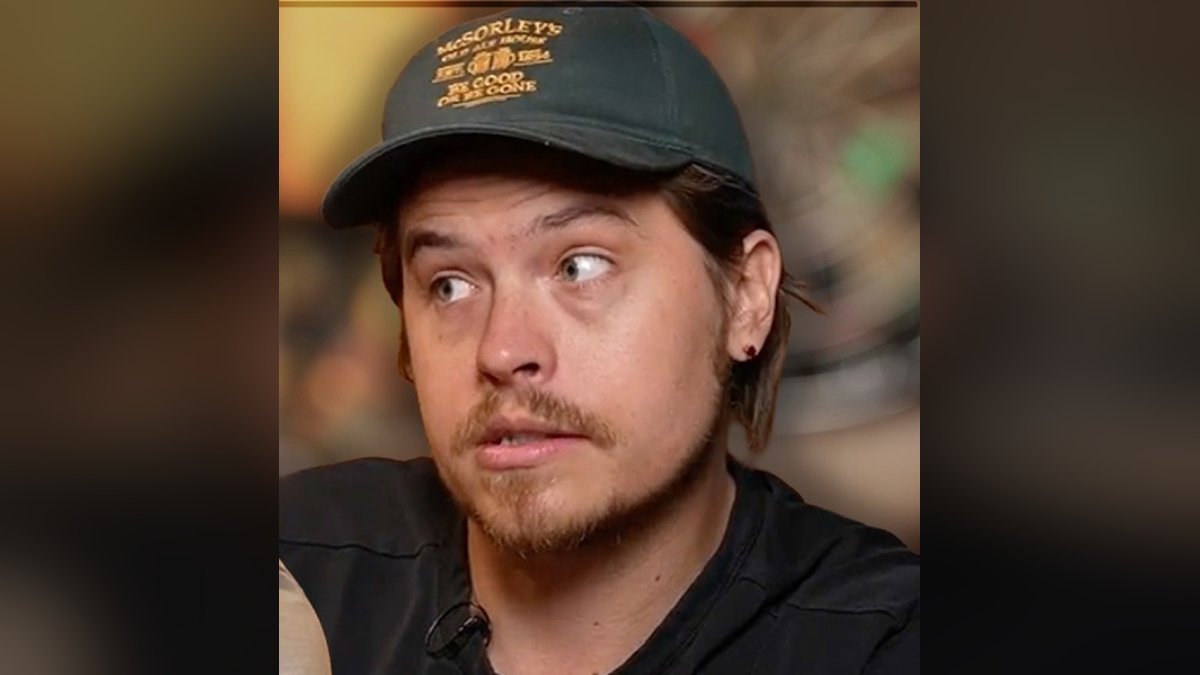 Dylan Sprouse in a dark green cap and black shirt glancing to the side during a podcast taping