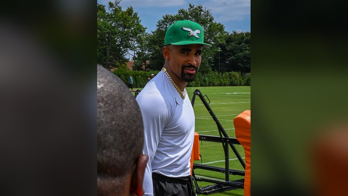 Eagles quarterback Jalen Hurts in a green Eagles cap and a white long-sleeve shirt stands at the edge of the practice field at training camp