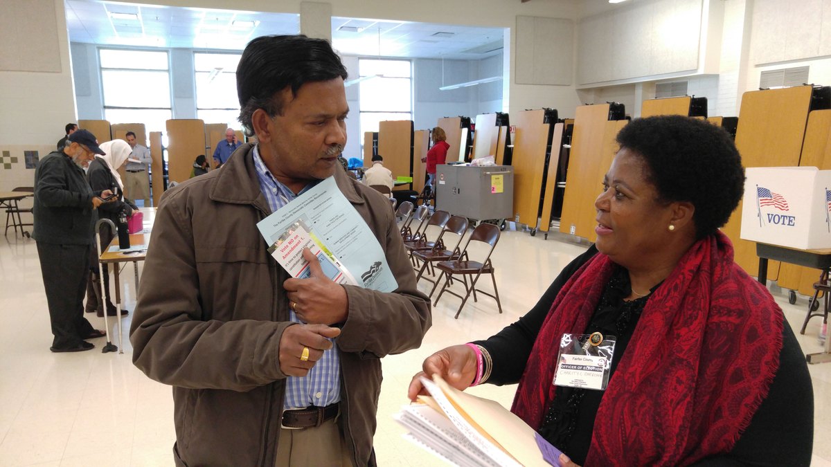 A voter holding a sample ballot talks with an election officer in a red scarf inside a school polling place lined with voting booths