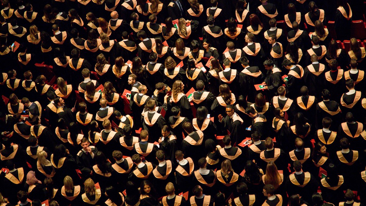 Hundreds of graduates in black caps and gowns seen from above filling an outdoor ceremony