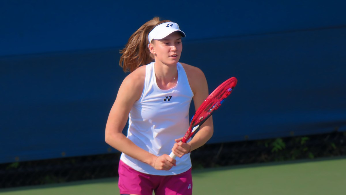 Elena Rybakina in a white top and visor holding her red racket on a hard court