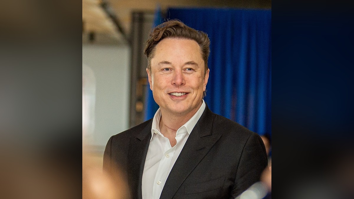 Elon Musk smiling in a dark blazer and white open-collar shirt in front of a blue curtain