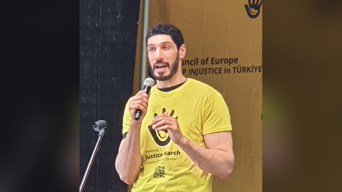 Enes Kanter Freedom in a yellow T-shirt speaking into a microphone on an outdoor stage