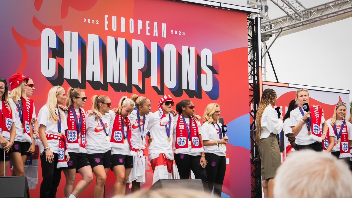 England women's players in white shirts and red and white scarves celebrate on a stage under a big European Champions sign