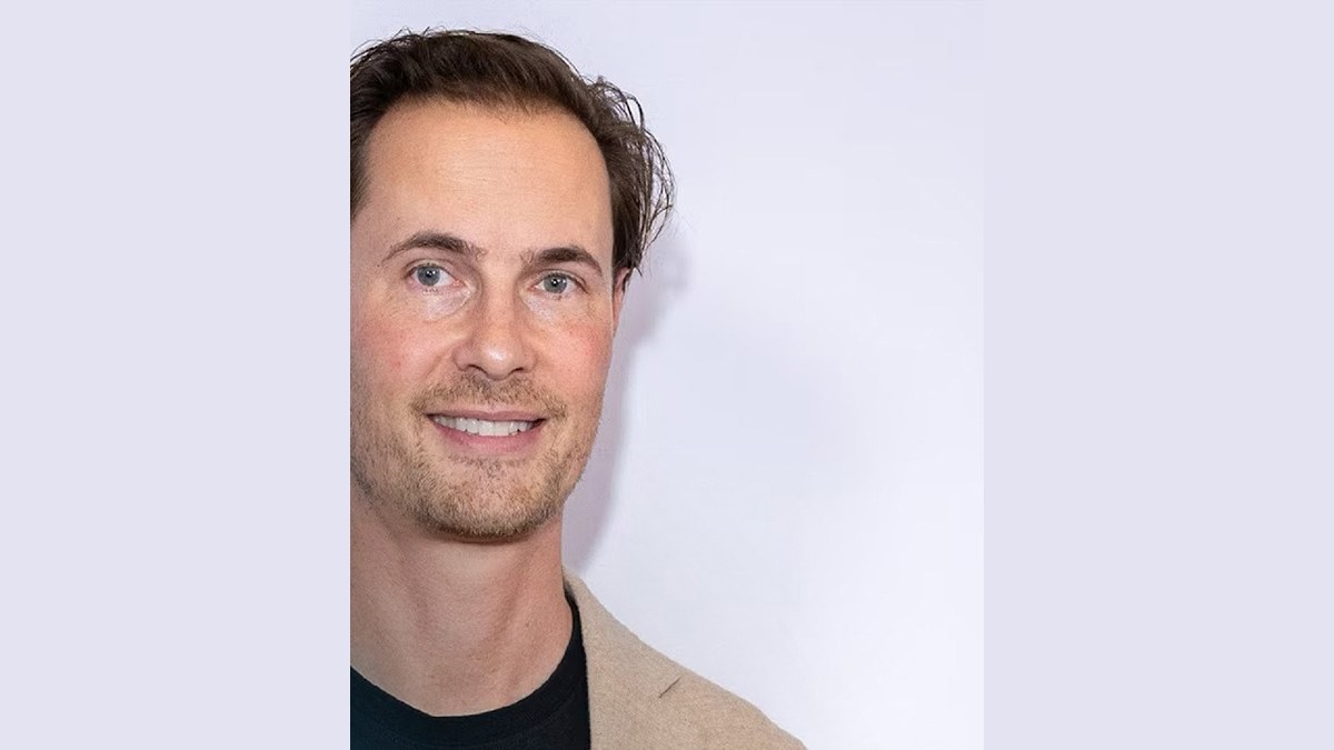 Erik von Detten smiling in a tan blazer and black T-shirt against a white backdrop