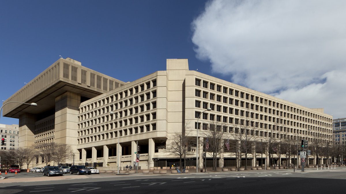 The tan concrete FBI headquarters building on a Washington street corner under a blue sky with a large white cloud