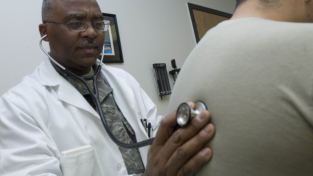 A doctor in a white coat holds a stethoscope to the back of a patient during an exam in a clinic