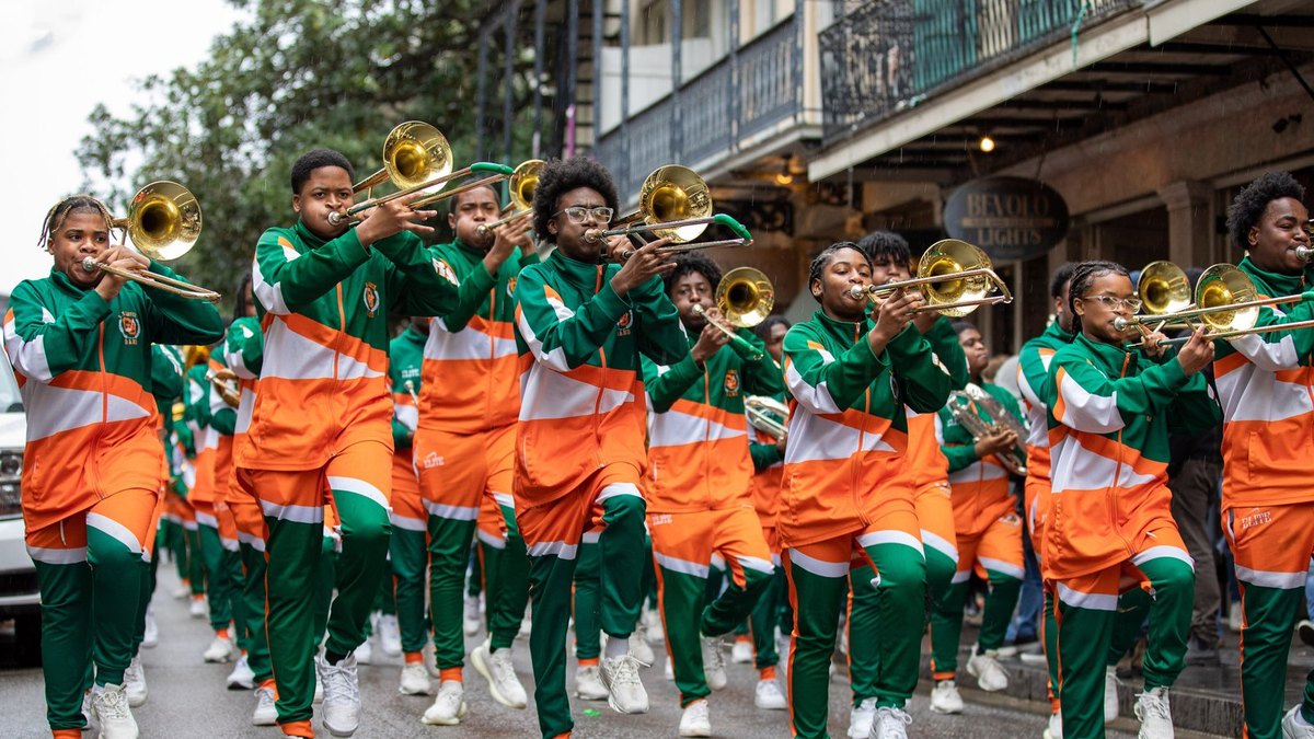 Young trombone players in bright orange and green uniforms march down a city street playing their horns during a parade