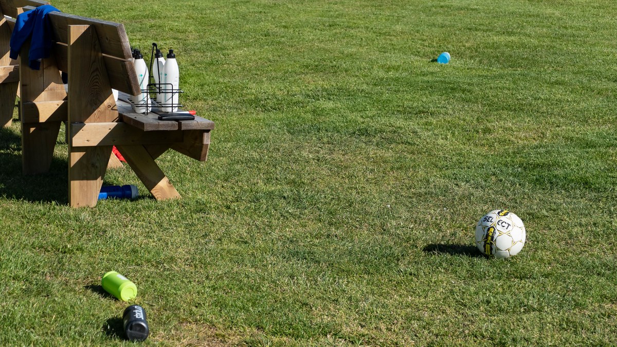 A soccer ball and scattered water bottles rest on the grass beside a wooden bench at an empty youth practice field