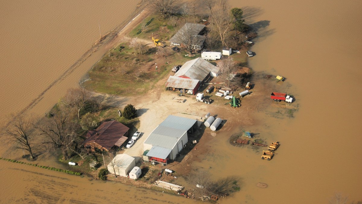 Aerial view of muddy brown floodwater surrounding a farm with barns, a house and parked trucks