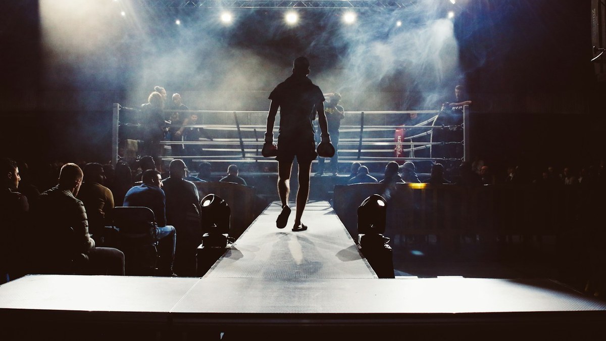 A fighter in silhouette walks down a lit ramp toward a boxing ring under bright arena lights and haze