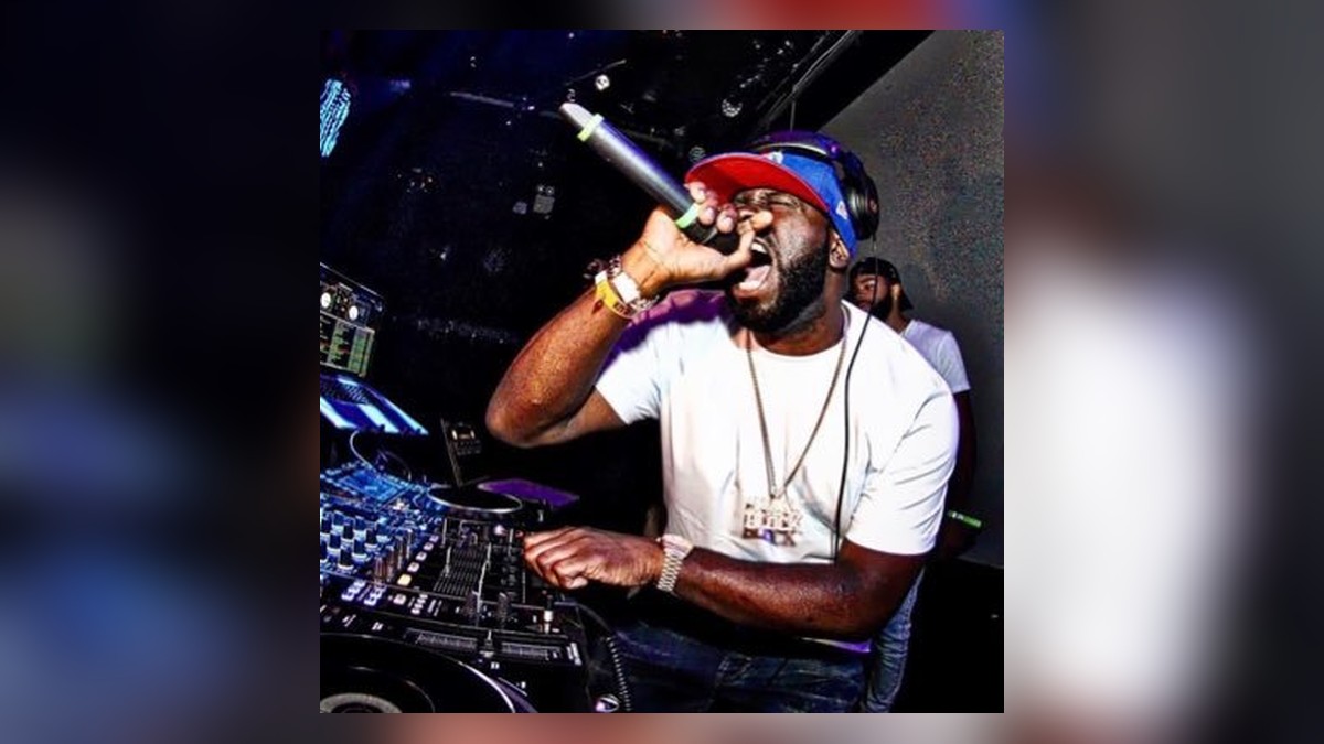 Funkmaster Flex shouting into a microphone behind DJ decks in a red cap and white tee