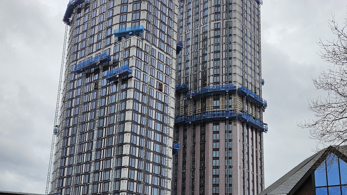 Two tall residential towers wrapped in blue construction panels rise against a gray sky during building work