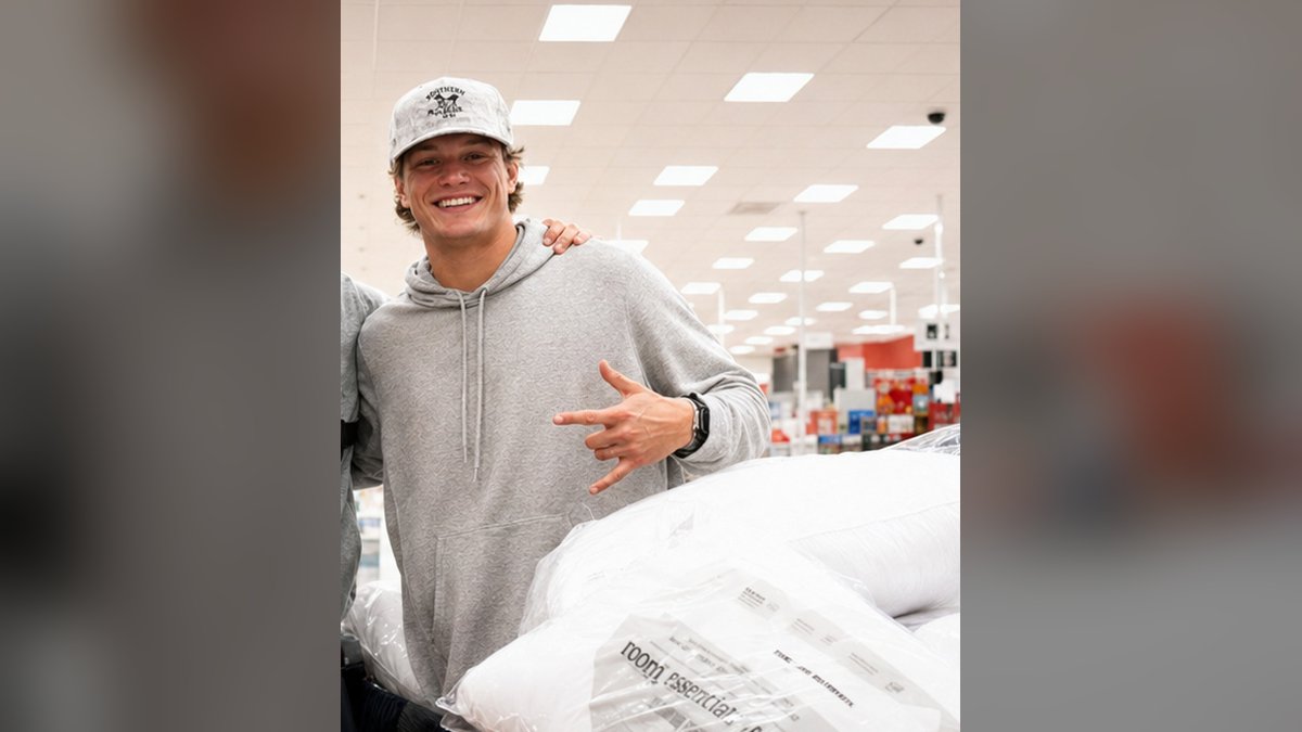 J.J. McCarthy smiles in a gray hoodie and white cap beside a cart full of pillows during a 2025 community shopping project