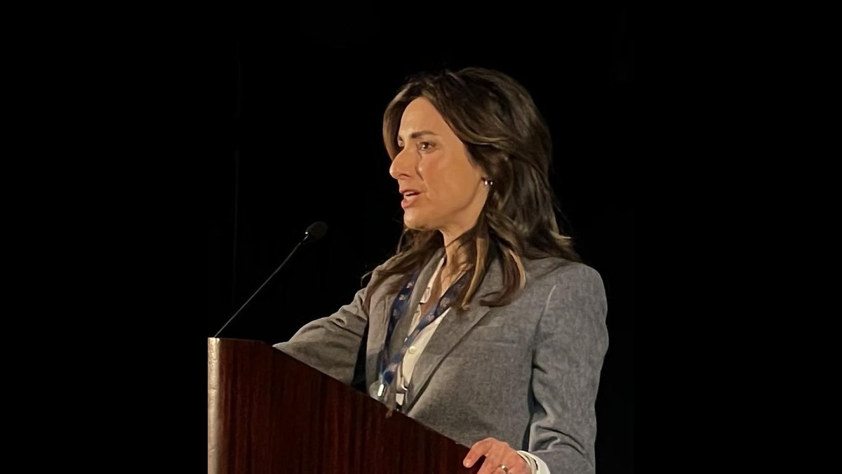 Gina Hinojosa in a gray blazer speaking at a wooden podium against a dark backdrop