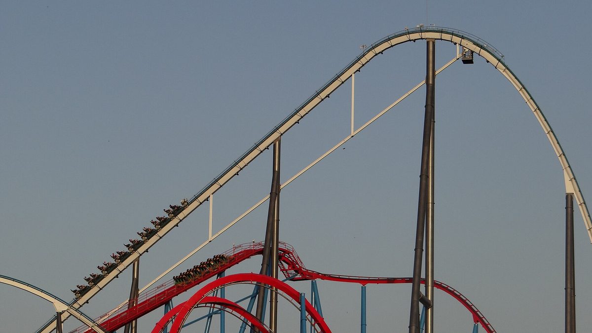 A tall white steel roller coaster lift hill rising above red looping track under a gray sky