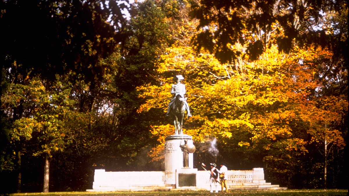 A bronze statue of a general on horseback stands on a stone monument under bright fall leaves at Guilford Courthouse battlefield