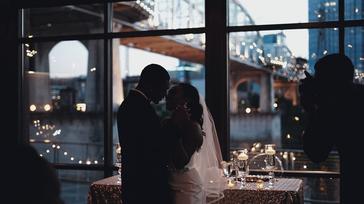 A bride and groom share a quiet dance in silhouette by tall windows as a guest films them against glowing city lights