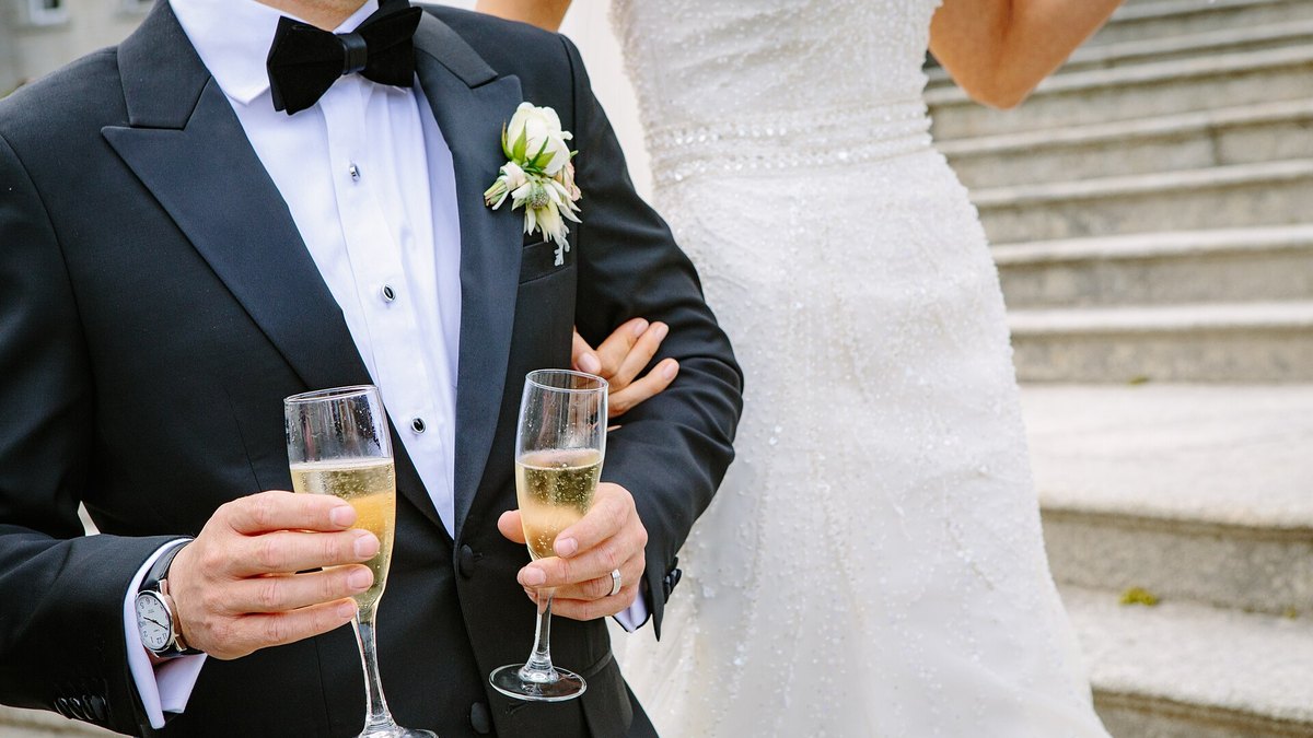 A groom in a black tuxedo and bow tie holds two glasses of champagne next to his bride in a white gown on stone steps