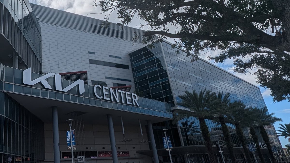 The Kia Center arena in downtown Orlando with its lettered sign above the entrance and palm trees along its glass facade