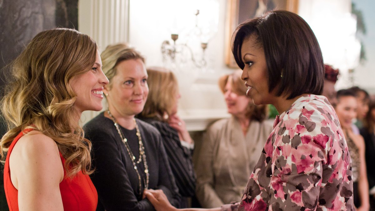 Hilary Swank in a red dress smiling as she greets Michelle Obama at the White House