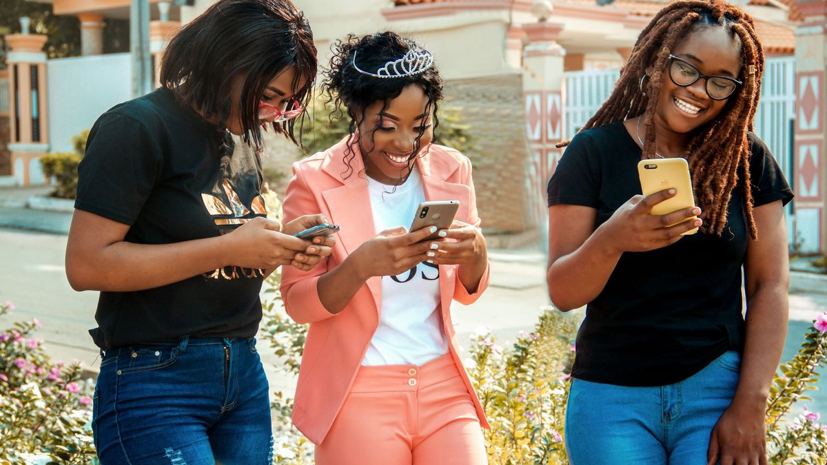 Three smiling Black women in stylish outfits looking at their phones outdoors with one wearing a pink suit and a tiara