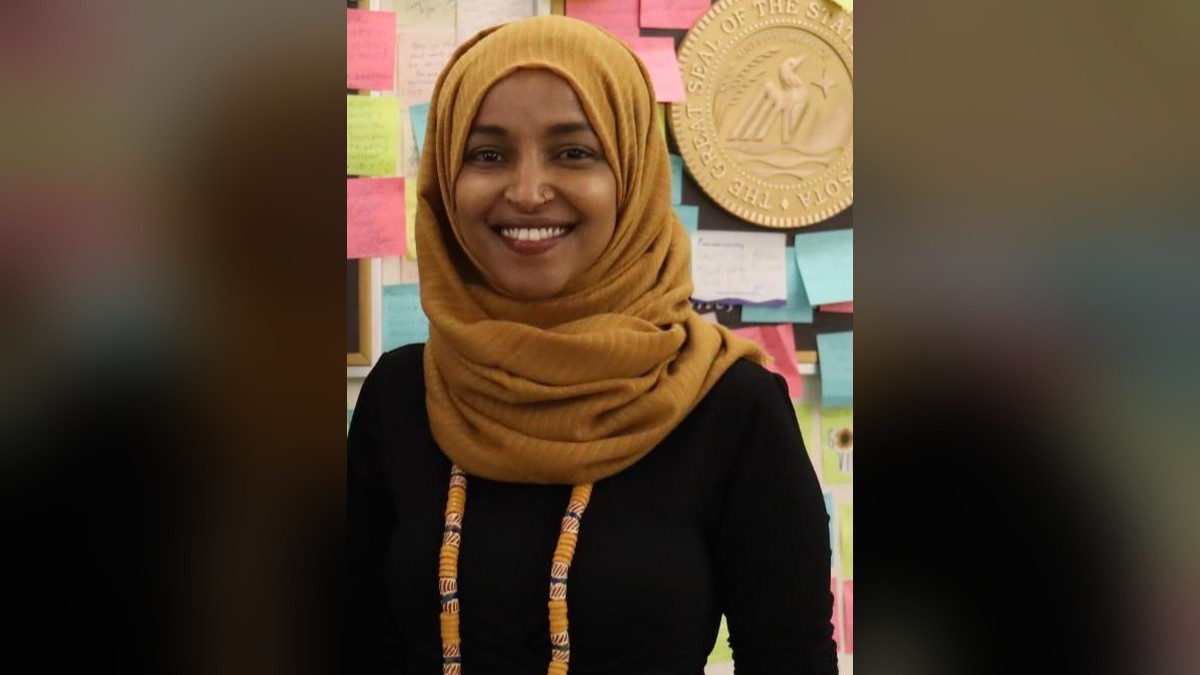 Rep. Ilhan Omar smiling in a mustard hijab and beaded necklace in front of a wall of colorful sticky notes