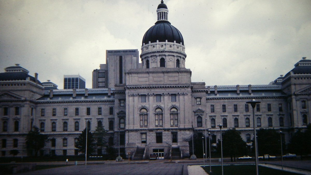 The dark dome and long gray stone front of the Indiana Statehouse in Indianapolis under a cloudy sky