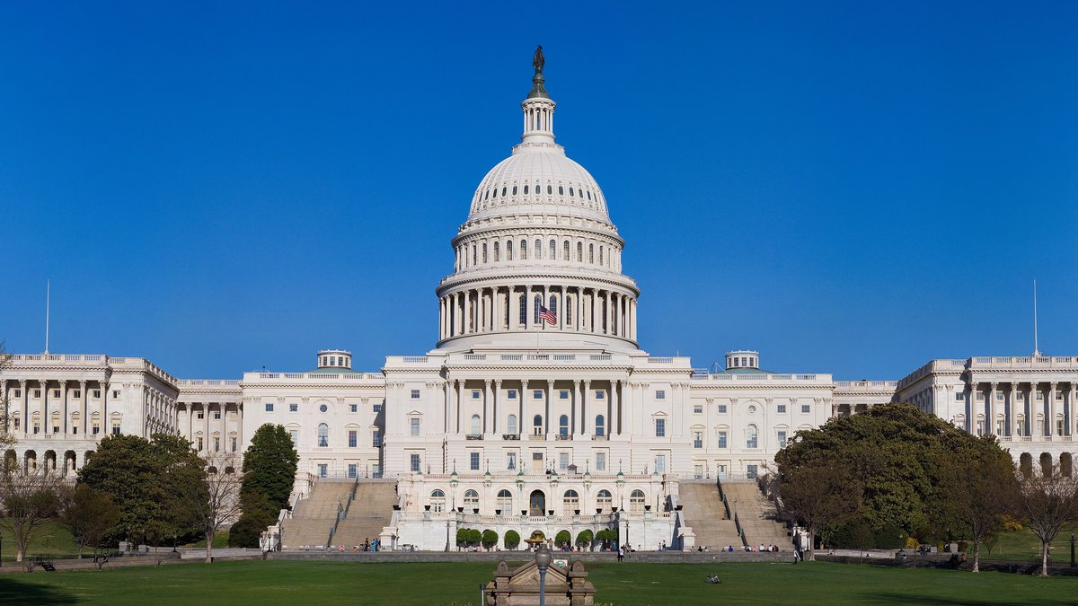 The west front of the United States Capitol with its white dome rising over green lawns under a clear blue sky