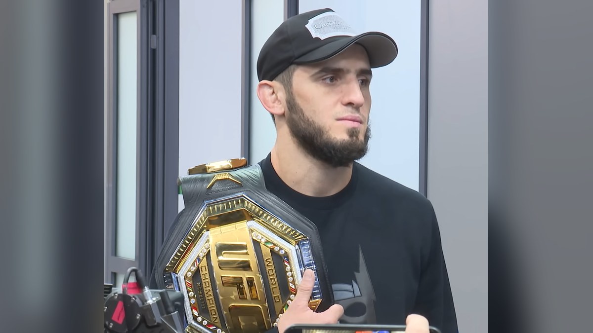 Islam Makhachev in a black cap and black T-shirt holding a gold UFC championship belt