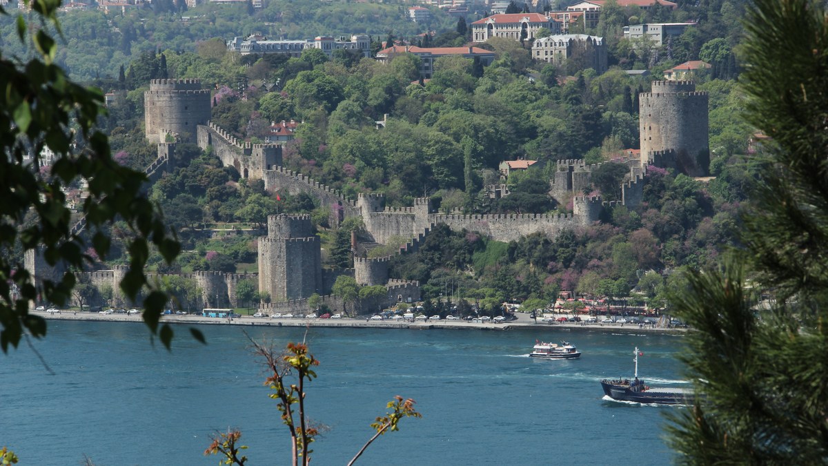 Stone towers and walls of a hillside fortress above the blue water of the Bosphorus with a ferry passing by in Istanbul