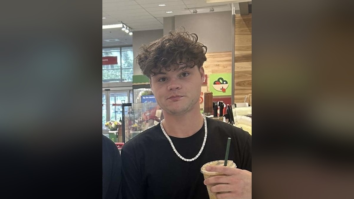 Jack Doherty with curly brown hair and a silver chain holding an iced coffee in a store
