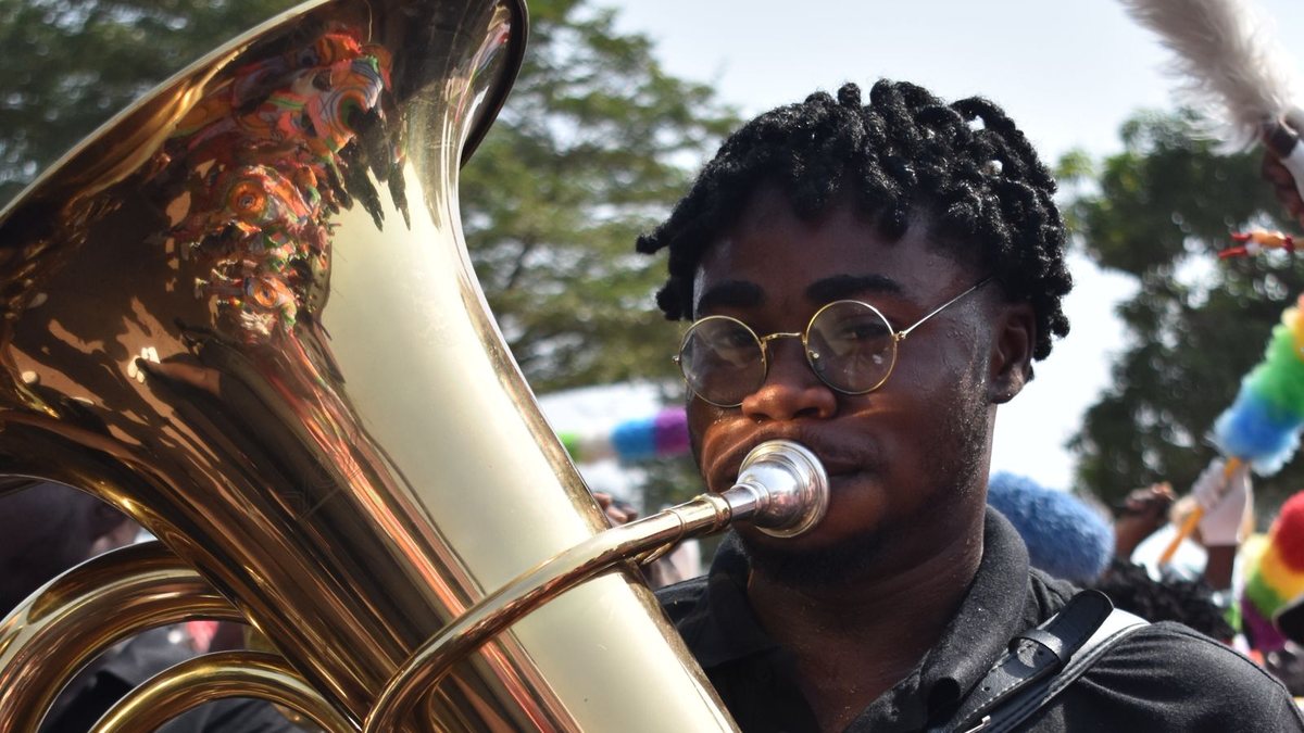 A young Black musician in round glasses playing a shiny brass tuba at an outdoor parade with colorful pom-poms in the background
