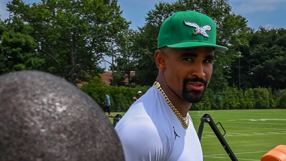 Eagles quarterback Jalen Hurts in a green Eagles cap and white shirt walks onto the practice field at training camp