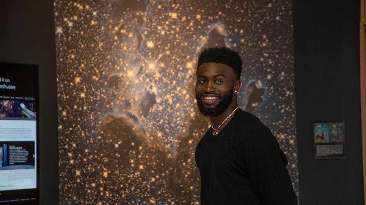 Jaylen Brown smiling in a black sweater in front of a glowing image of stars and space clouds