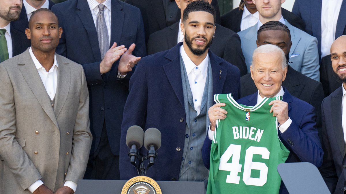 Boston Celtics star Jayson Tatum in a navy suit next to a green Celtics jersey at the team's 2024 championship celebration at the White House
