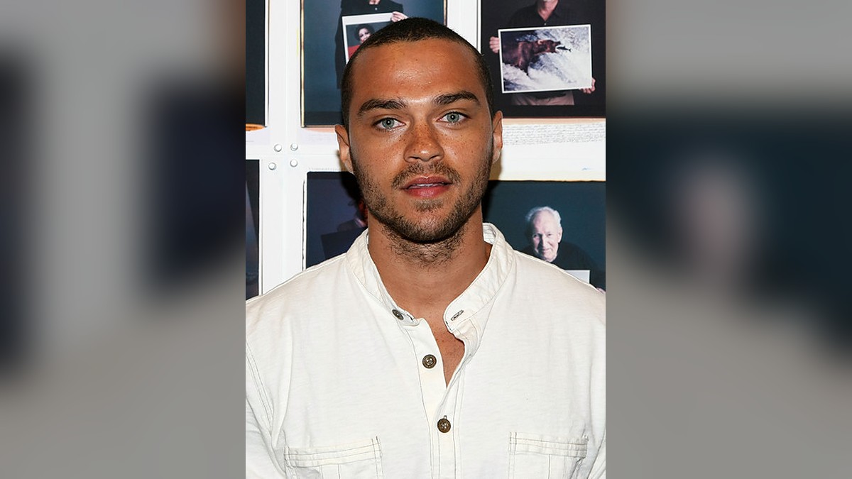 Jesse Williams in a white button-front shirt standing in front of a wall of framed portrait photographs