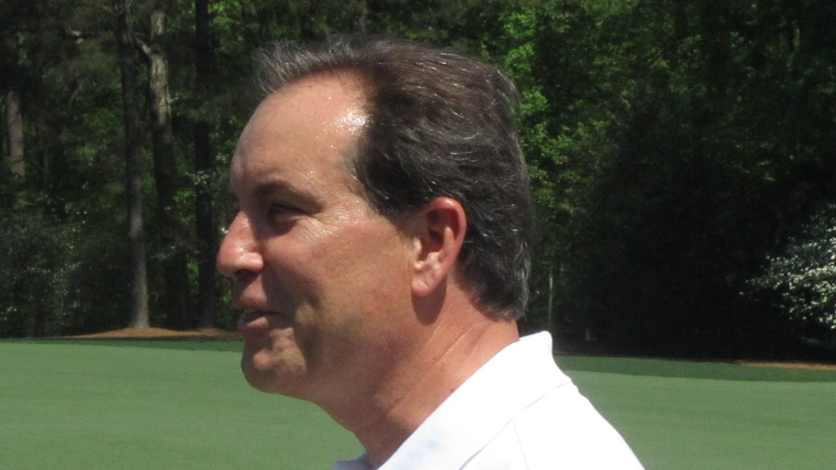 Jim Nantz in a white polo shirt smiles in profile on a sunny golf course lined with tall pine trees