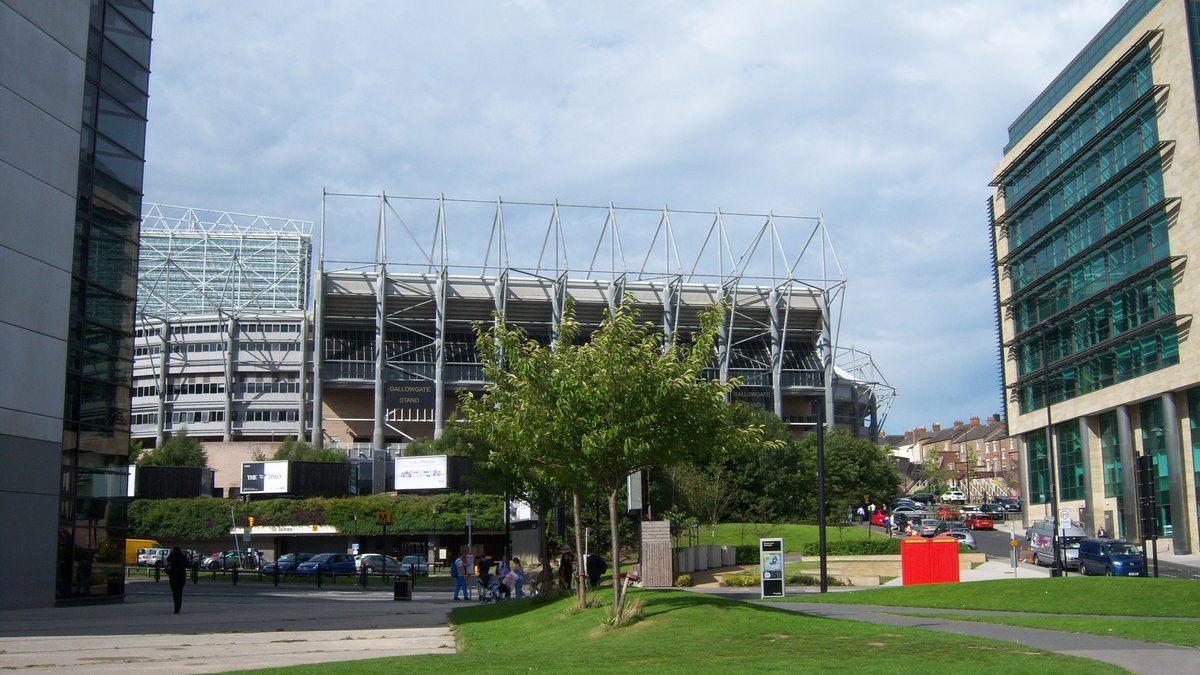The tall steel roof supports and stands of St James' Park rise above trees and a green lawn in Newcastle under a cloudy sky