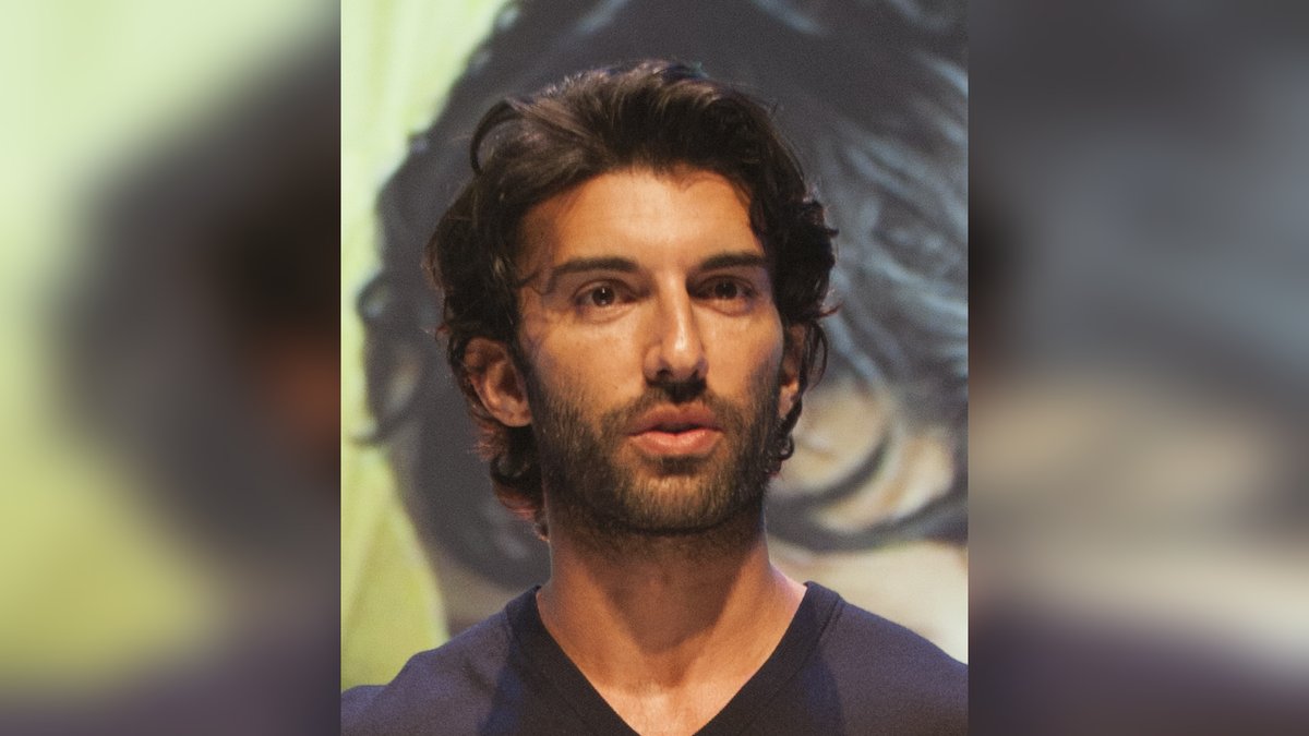 Justin Baldoni with dark wavy hair and a beard looking toward the camera in a dark V-neck shirt