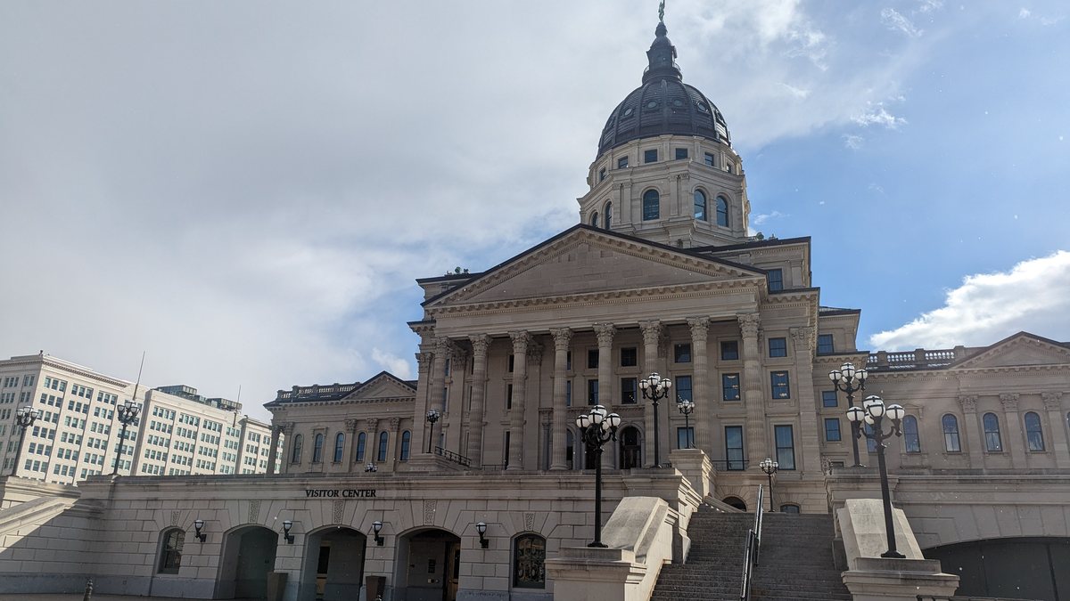 The domed Kansas State Capitol in Topeka with its columned entrance and stone plaza under a partly cloudy sky