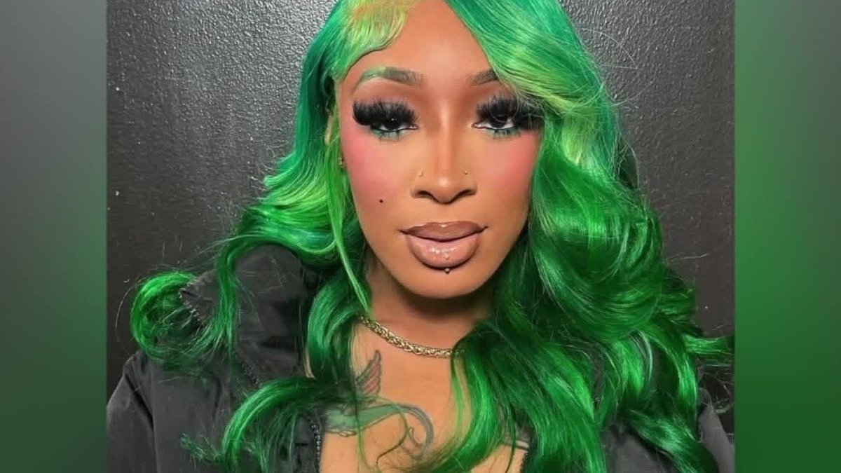 Kash with bright green wavy hair in a black jacket against a dark backdrop