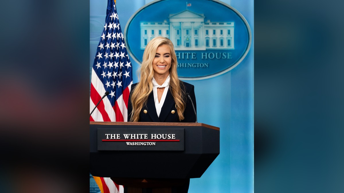 Katie Zacharia smiling in a navy blazer at the White House briefing room podium beside an American flag