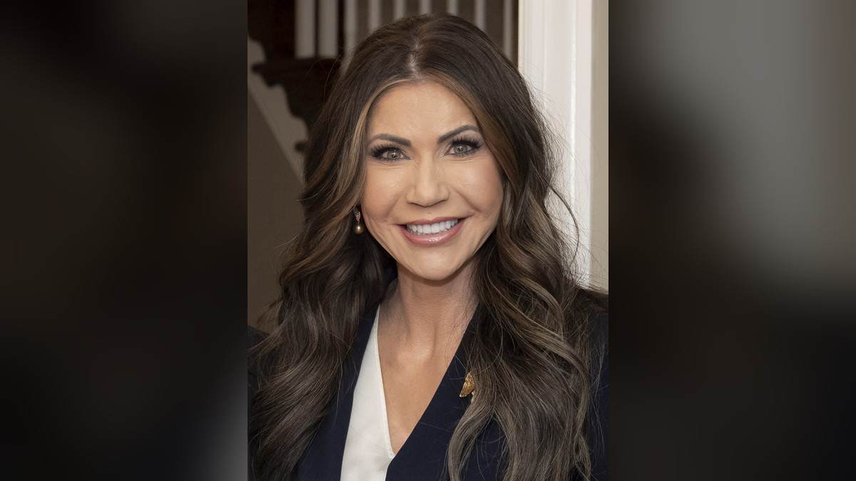 Kristi Noem smiling with long dark wavy hair in a navy blazer and white blouse