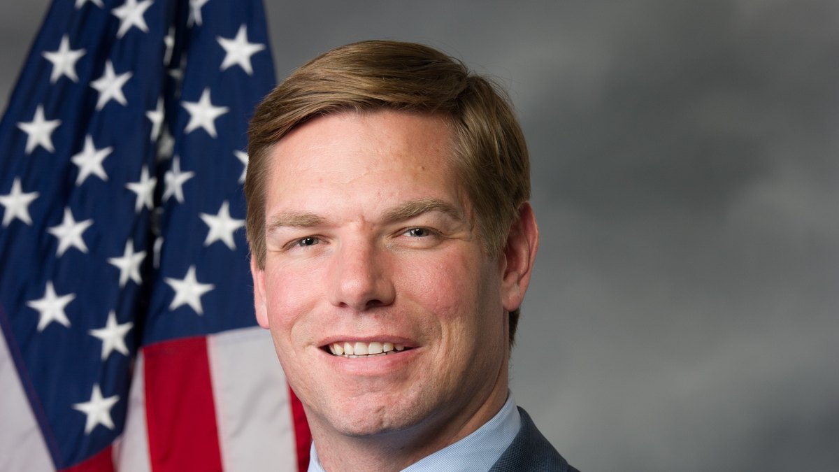 Official congressional portrait of Eric Swalwell smiling in a blue suit and tie beside an American flag