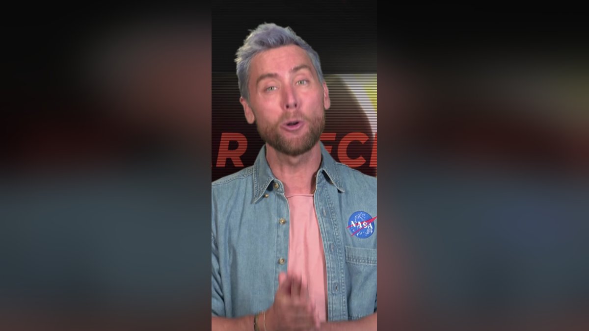 Lance Bass in a denim shirt speaking to the camera in a NASA studio
